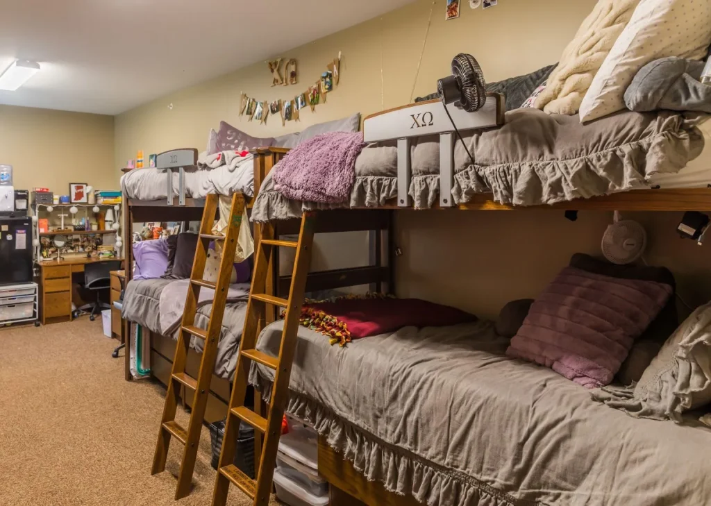 Four bunk beds with gray bedding in a cozy student housing dorm room, decorated with pillows, photos, and string lights.