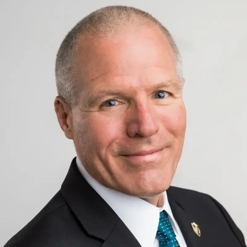 Smiling man in a suit and teal tie with short gray hair, posing against a light gray background, embodying professionalism and confidence in leadership & governance.