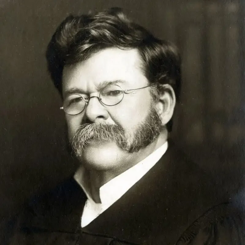 Black-and-white portrait of a man with glasses, large mustache, and mutton chops, wearing a dark coat—a distinguished figure reflecting leadership & governance.
