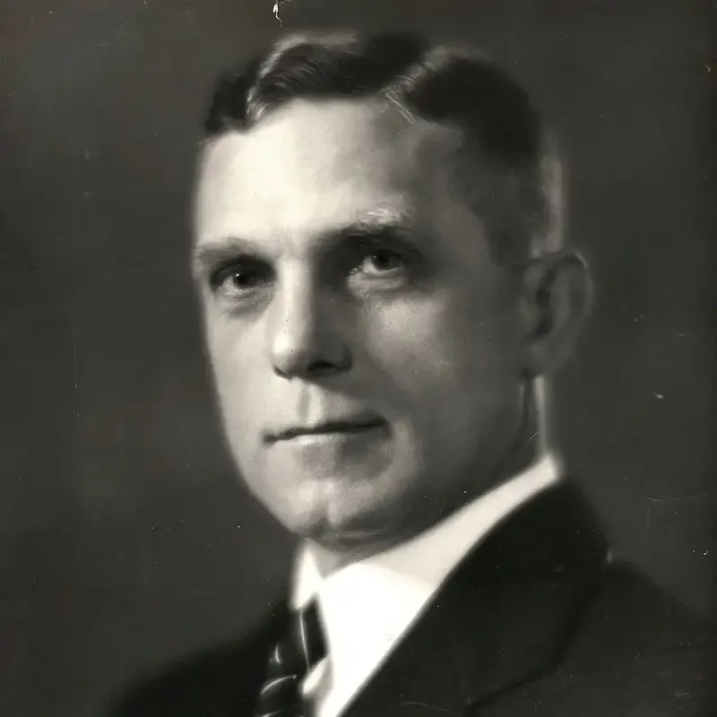 Black and white portrait of a man in a suit and tie, facing slightly to the left with a neutral expression, embodying an air of leadership and governance.