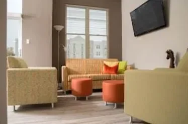 Modern living room at Promenade Apartments with beige chairs, orange ottomans, a sofa, and a wall-mounted TV near a large window.