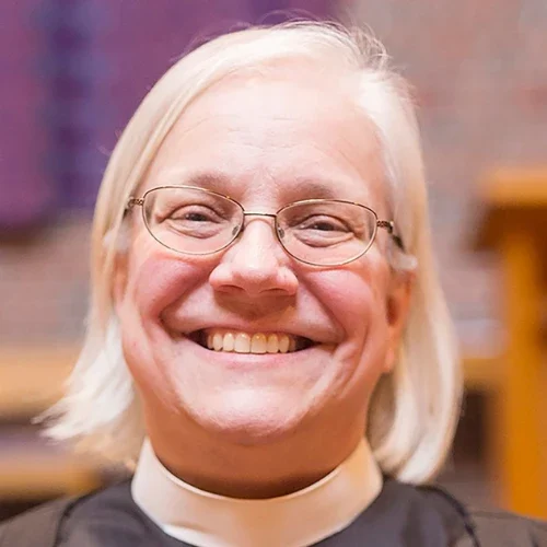Smiling person with glasses and white hair wearing a clerical collar, indoors with blurred background, reflecting Our Lutheran Identity.