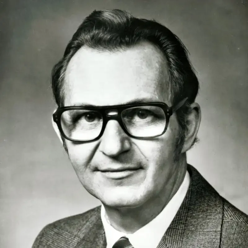 Black and white portrait of an older man wearing glasses, a suit jacket, and a tie, smiling slightly—exuding wisdom and experience in leadership & governance.