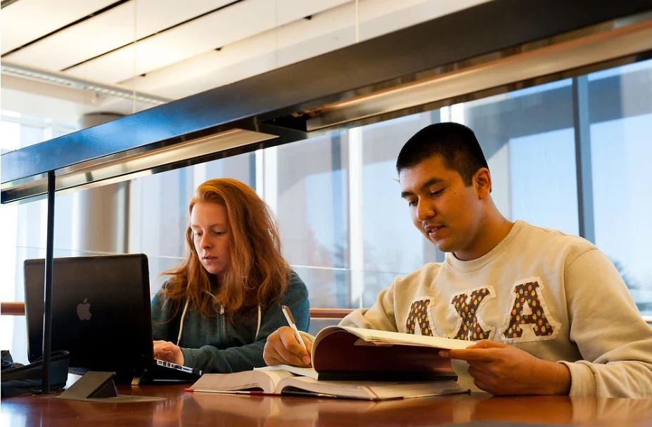Two students studying at a Vale table, one using a laptop and the other writing in a book.