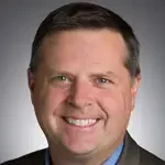 Smiling man in a suit jacket and blue shirt, with short brown hair, exuding confidence in communications, against a gray background.