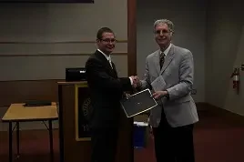 Two men in suits shake hands while one receives an engineering certificate during an indoor award ceremony.