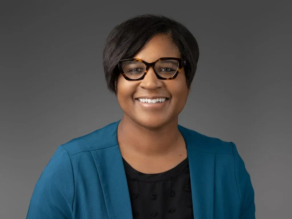 Smiling woman with short dark hair, glasses, blue blazer, and black top on a gray background.