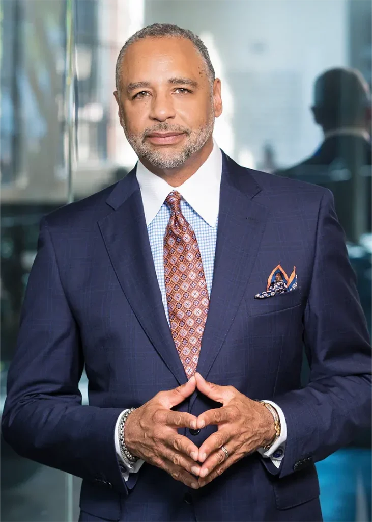 A confident man in a navy suit stands indoors, hands clasped, smiling at the camera.