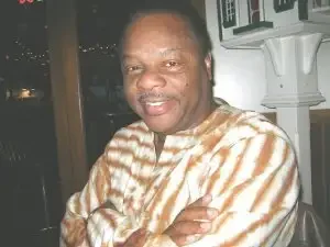 A man in a striped shirt sits indoors with arms crossed, smiling at the camera.