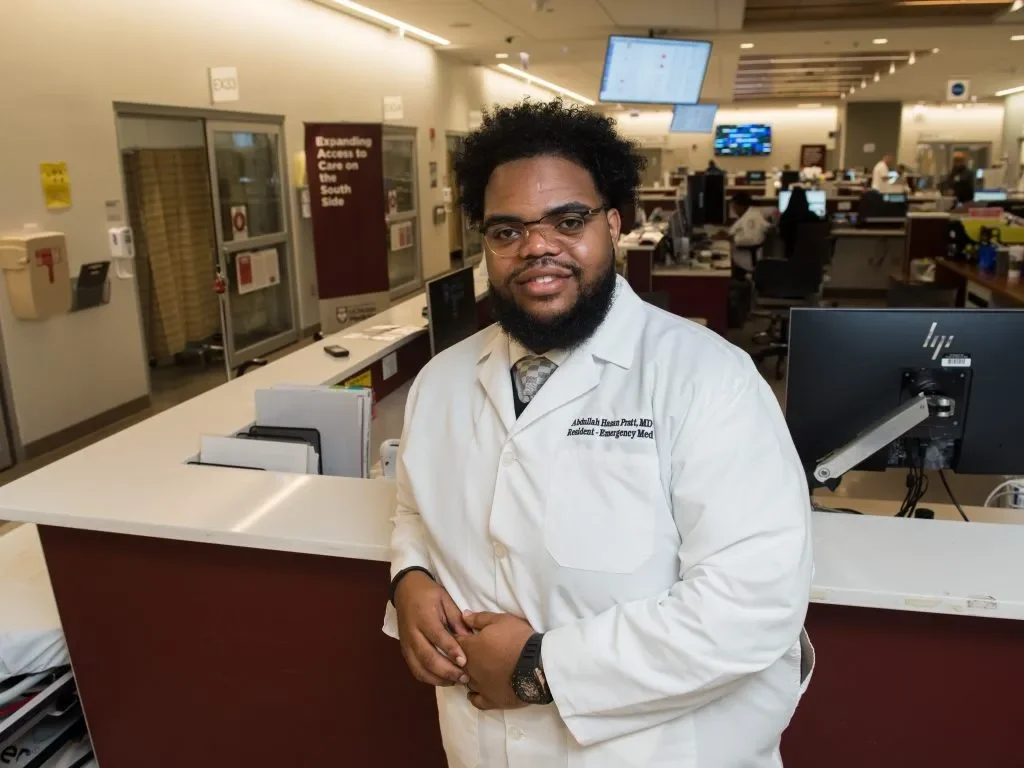 A doctor in a white coat stands smiling at a hospital reception desk with computers in the background.