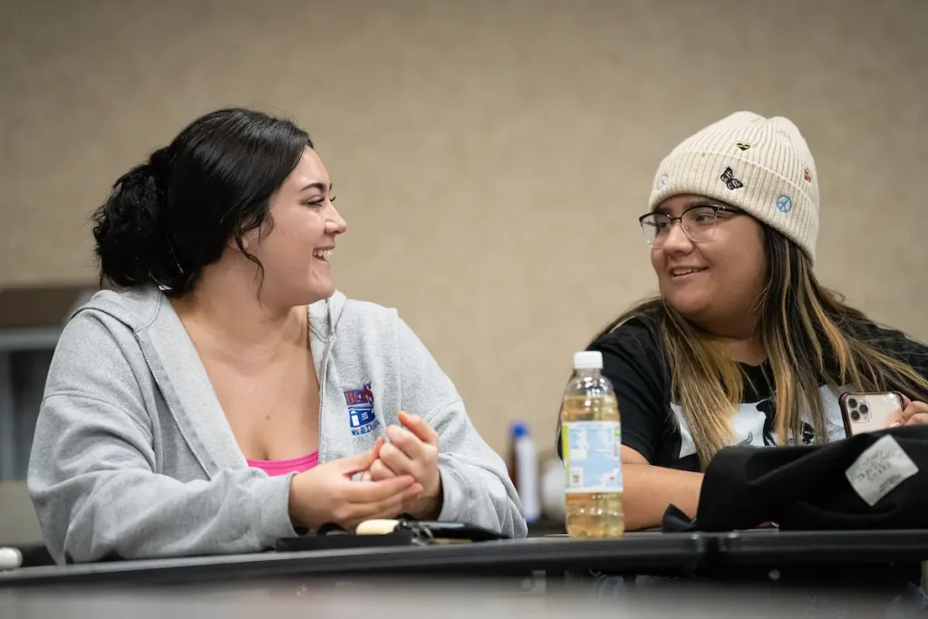 Two young women sit at a table in Vale, smiling and talking; one wears a beanie and glasses, the other a gray hoodie.