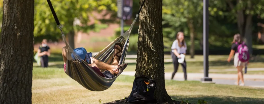 A student relaxes in a hammock between trees on campus, enjoying the day and demonstrating positive student behavior, while peers walk by in the background.