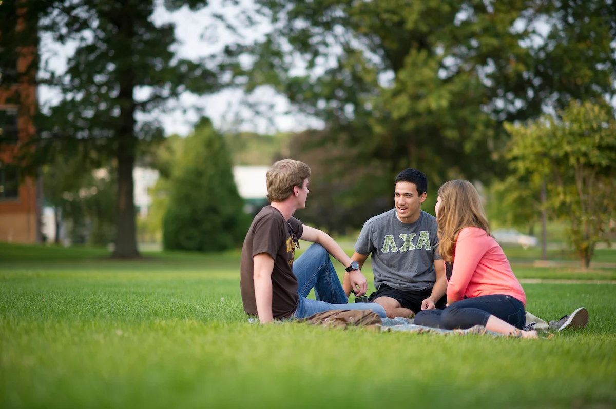 Three young adults sit on a blanket in the grass, talking and relaxing in a park-like setting, enjoying time together and chatting about life.