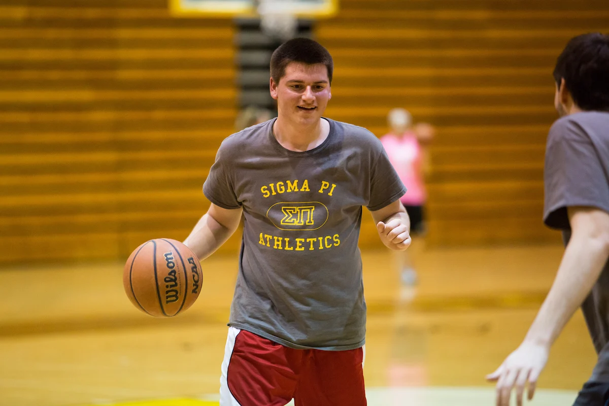 Young man dribbling a basketball indoors on campus, wearing a “Sigma Pi Athletics” shirt and red shorts.