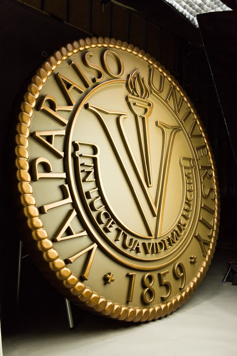 Large gold Valparaiso University seal with a torch, "1859," and Latin motto displayed on a round plaque in the President's Office.