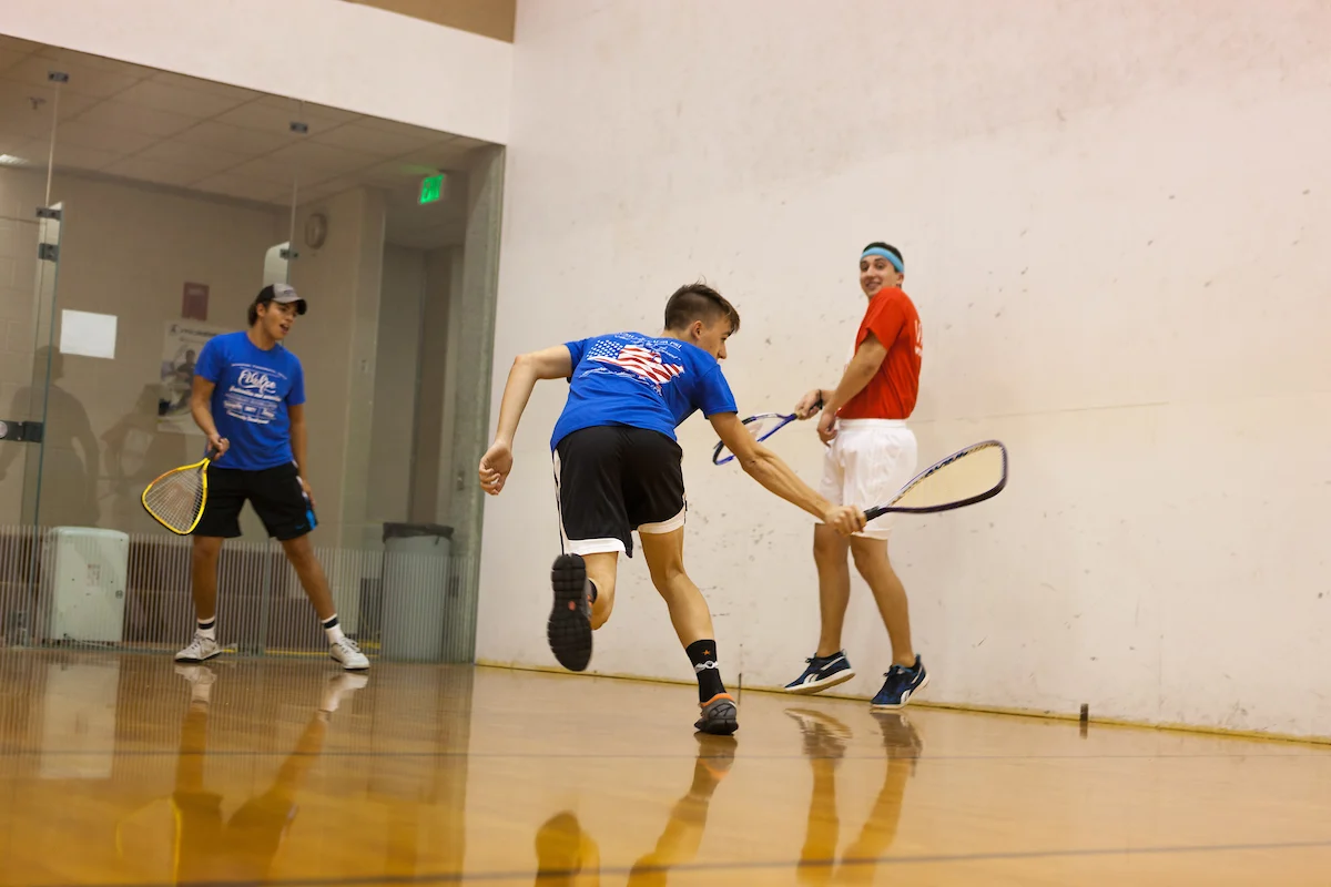 Three people playing racquetball on a campus indoor court, with one player swinging at the ball.