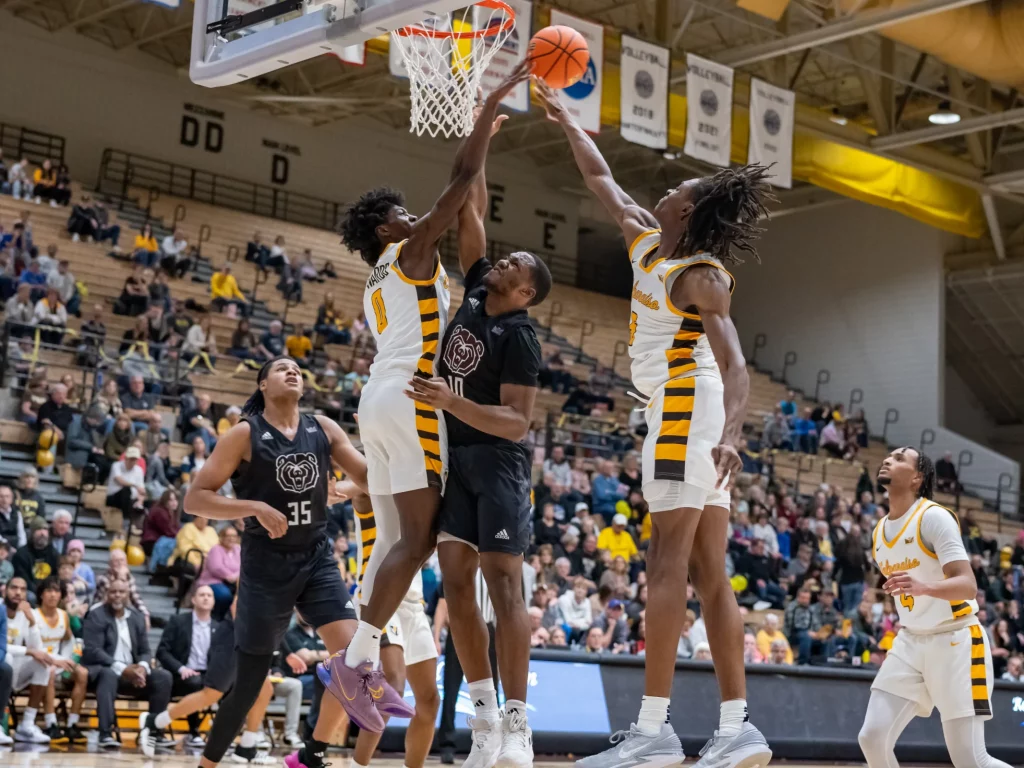 Two basketball teams compete as players jump to block a shot near the hoop during a game in a gymnasium.