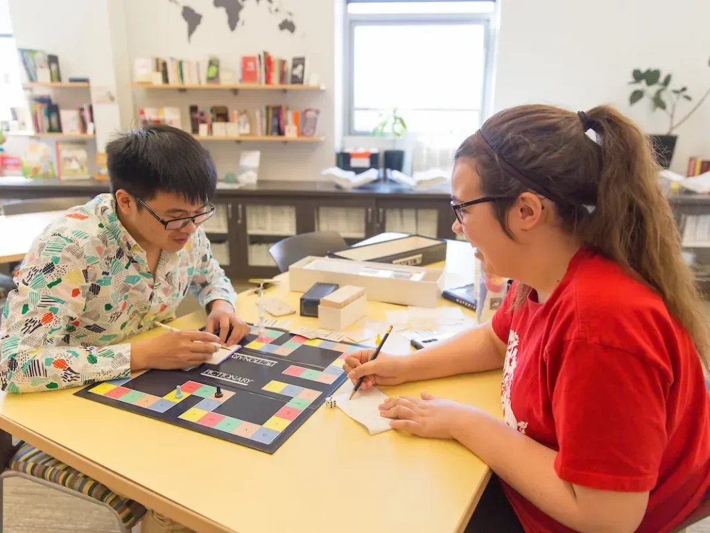 Two people prep for study abroad as they play a board game together at a table in a bright, cozy room with bookshelves and a world map.