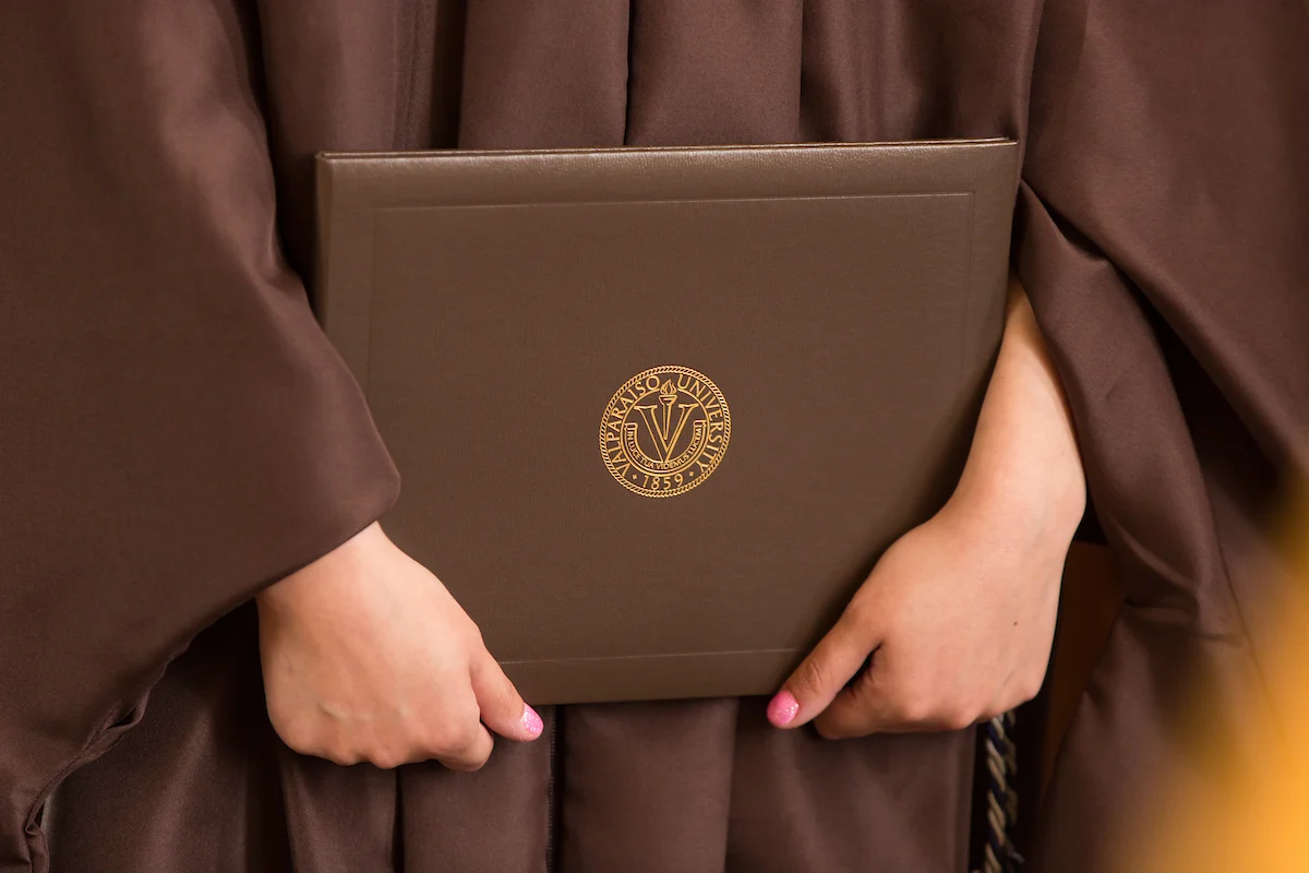 Person in graduation robe holding a brown diploma folder with a gold university seal on the cover, celebrating a proud milestone. Information for Graduates is available to help guide you through this memorable achievement.