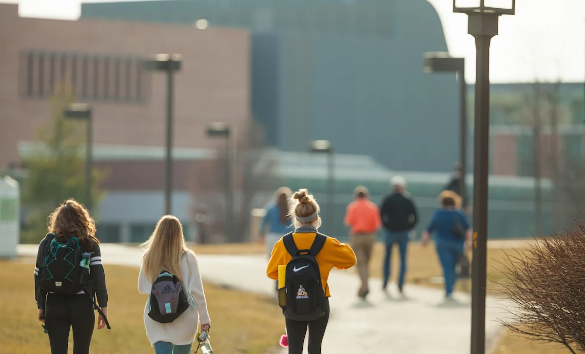 Students with backpacks walk on a campus pathway toward academic buildings on a sunny day, ready for classes and assessment.