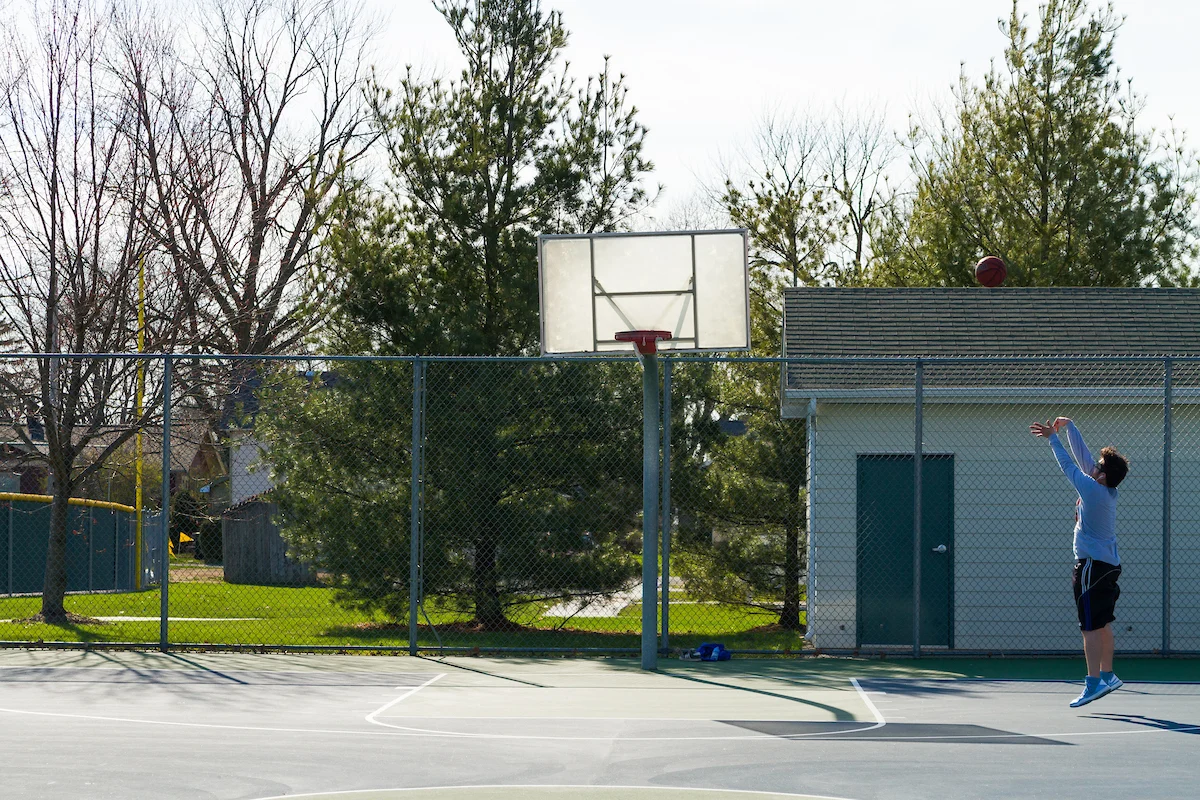 A person shooting a basketball toward a hoop on an outdoor court, with trees and a building in the background, demonstrates their assessment of distance and aim.