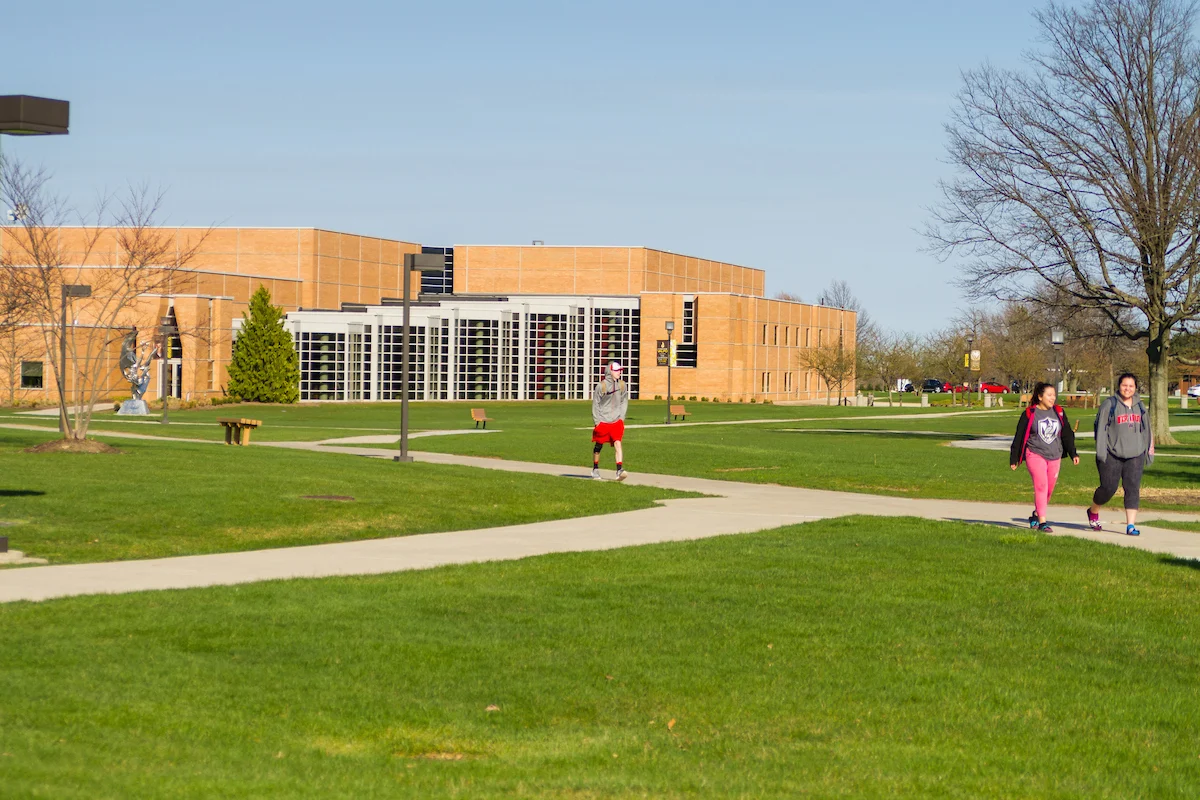 Three people walk on a sidewalk in front of a large brick building, perhaps heading to an assessment, on a sunny day with green grass and trees.