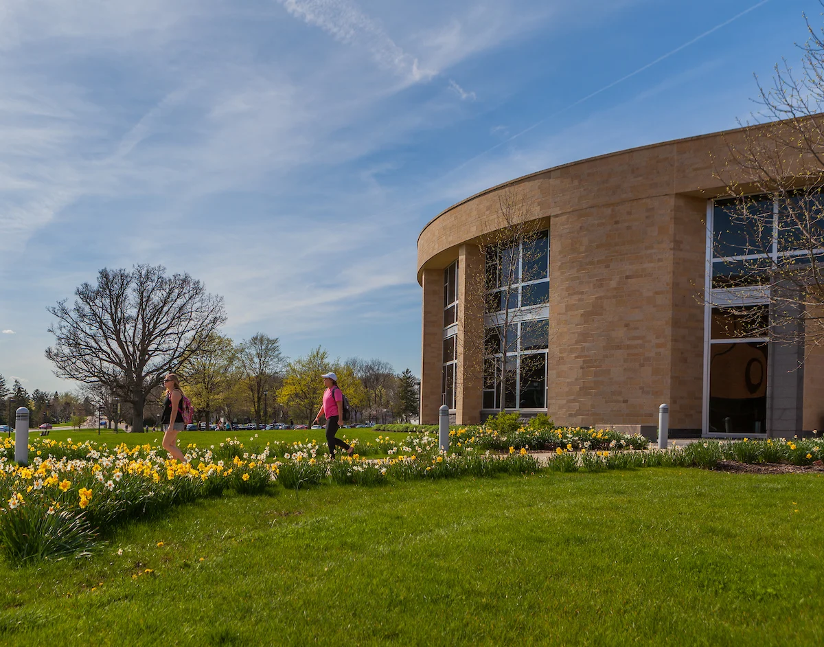 Two people walk past blooming flowers near the modern Center for the Arts building on a sunny day.