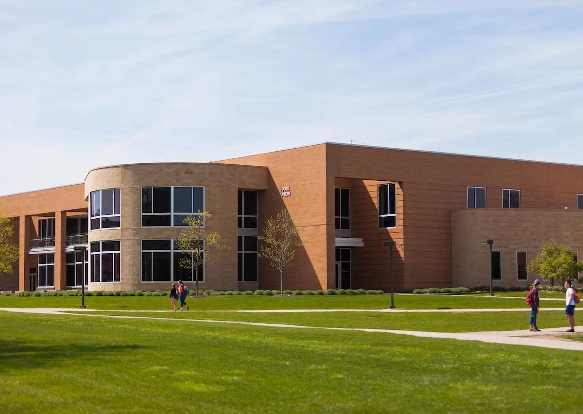 A modern brick and glass campus building stands tall as students cross the green lawn, heading to their next class or assessment.