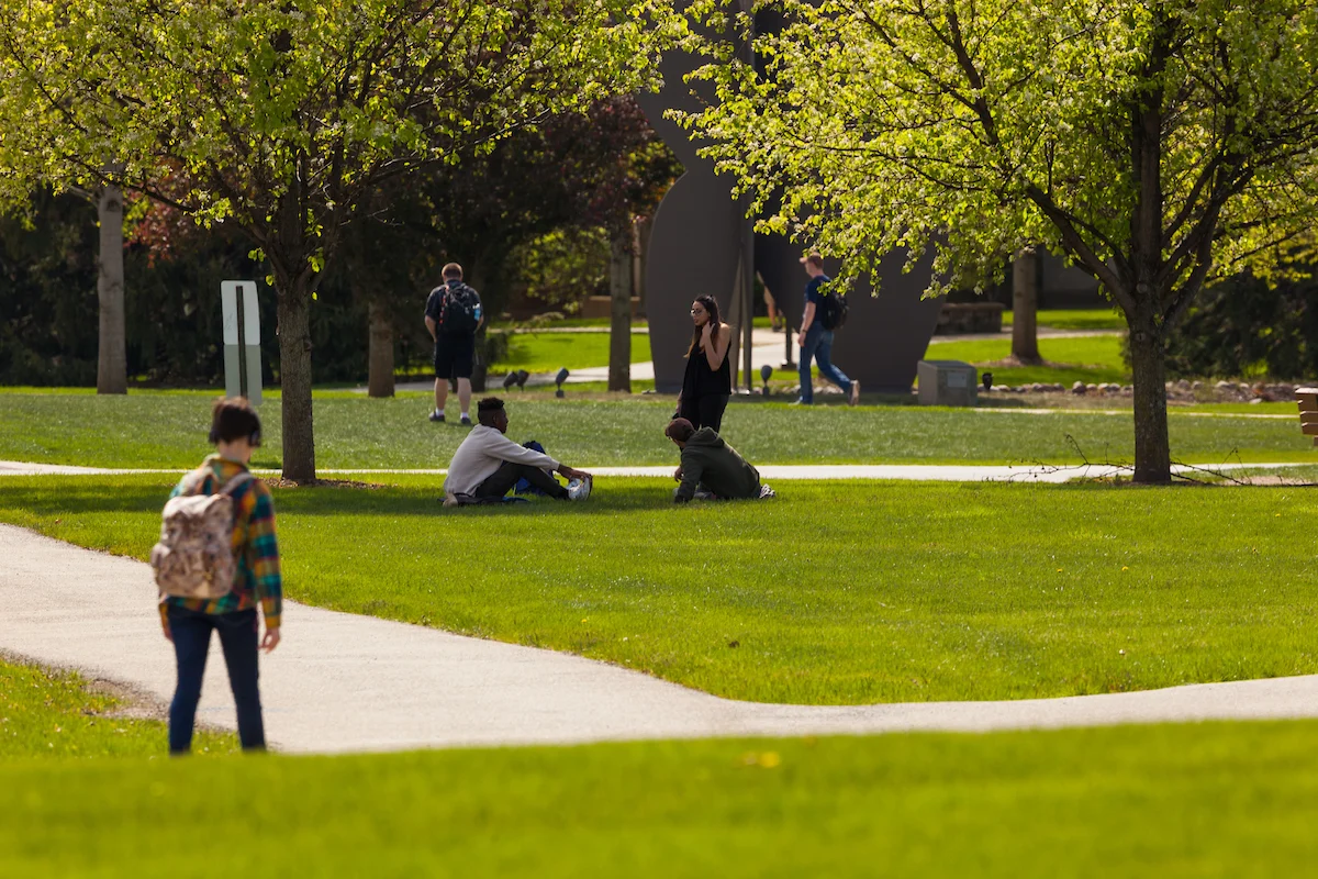 Students sitting and walking on a green, tree-lined college campus on a sunny day, using Maps & Directions to explore and find their next class.