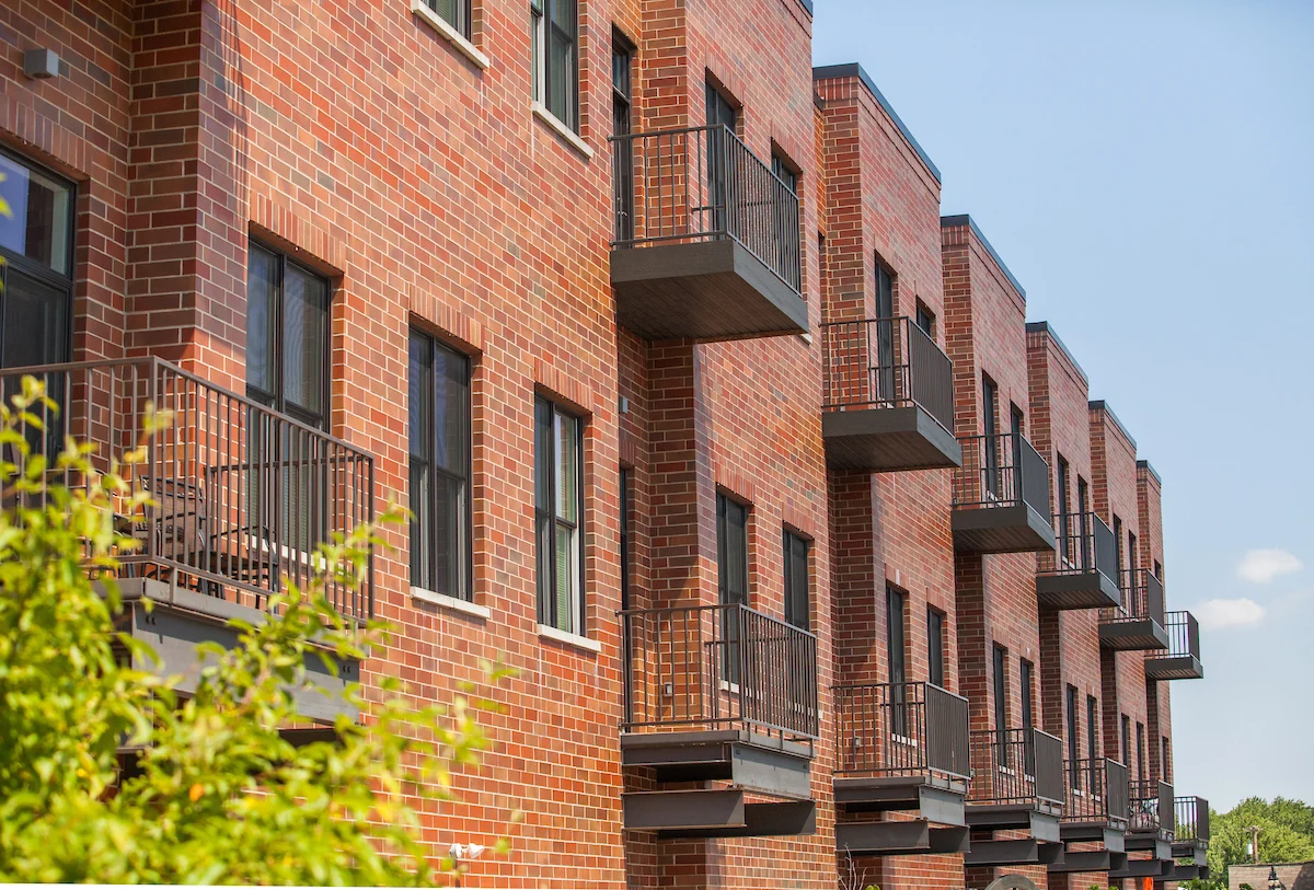 Promenade Apartments is a red brick apartment building featuring several small black metal balconies, set against a clear blue sky.
