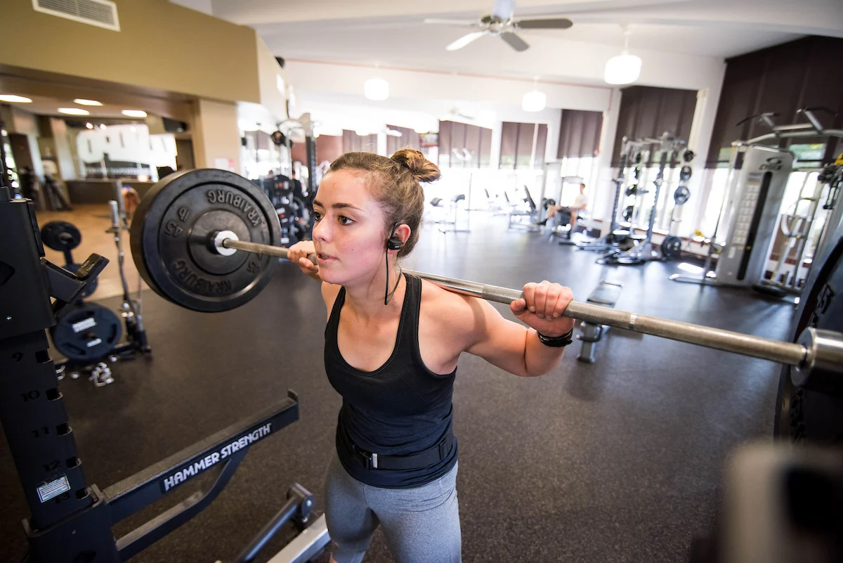 Woman in gym squatting with a barbell on her shoulders, wearing workout clothes and earbuds on a bustling campus.