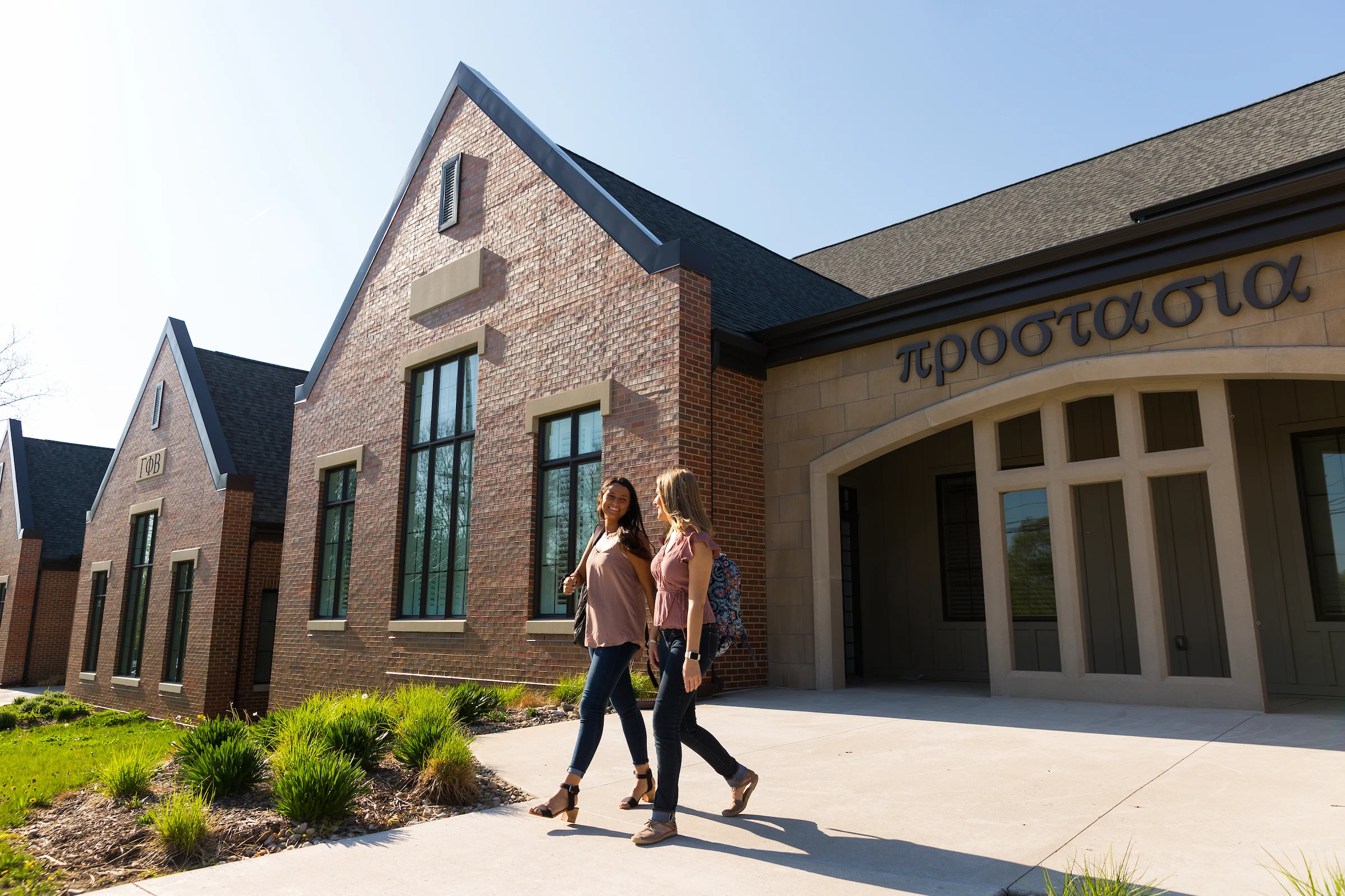 Two women walk and talk outside a brick housing building with large windows and Greek letters above the entrance.
