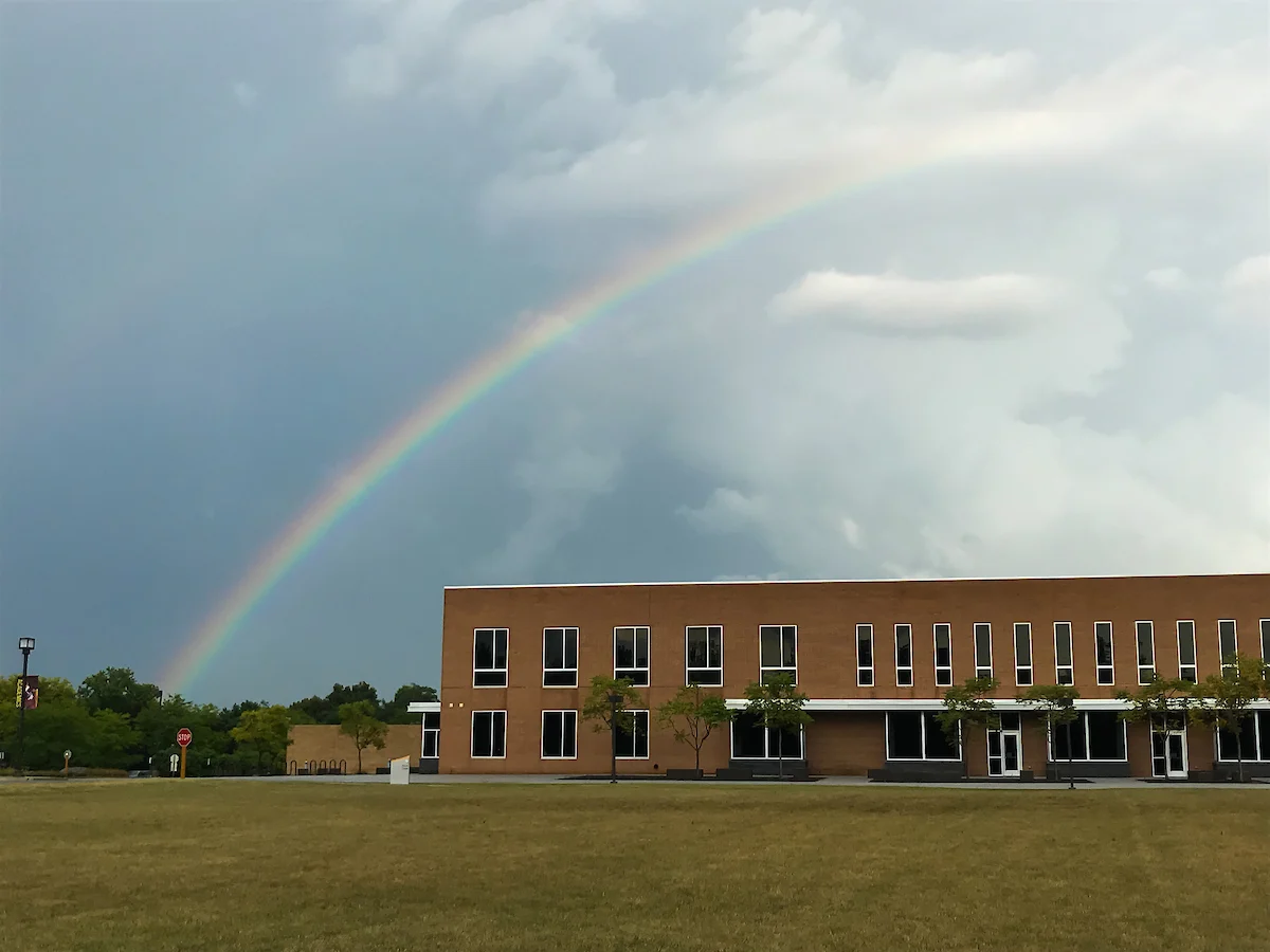 A rainbow arches over a brick building and grassy field under a cloudy sky, symbolizing hope and the promise of multicultural resources within the community.