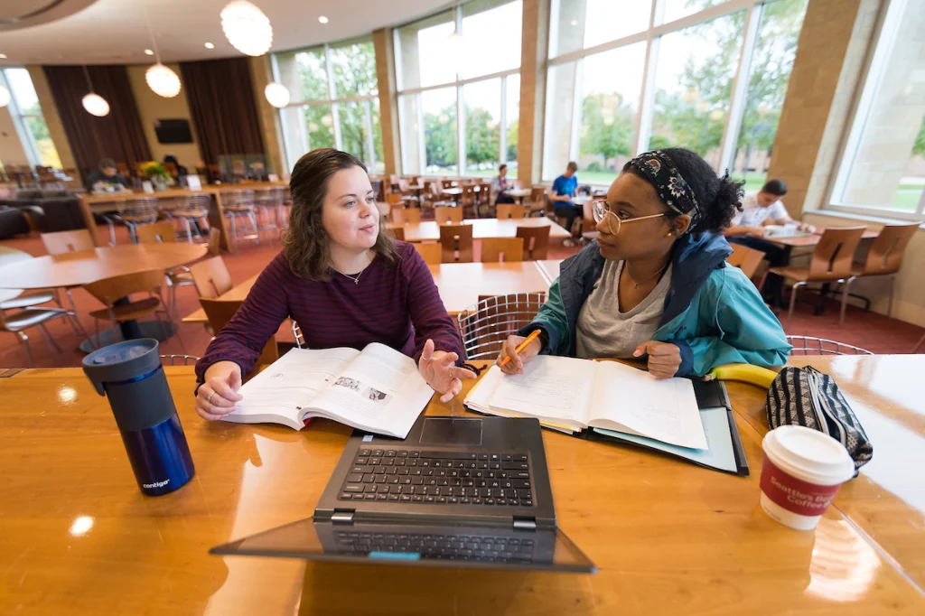 Two students study together at a table with books, notebooks, and a laptop in a bright, modern cafeteria.