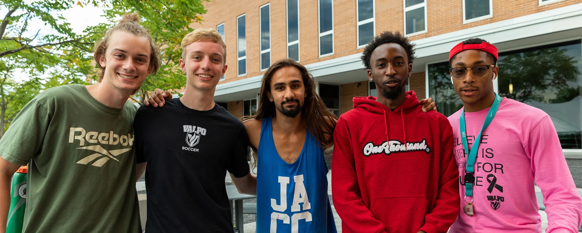 Five young men stand together outdoors during Welcome Week, smiling with a building and trees in the background.