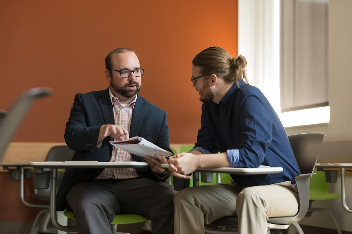Two men sit at desks talking in a modern Vale classroom with orange walls, one holding papers and gesturing.