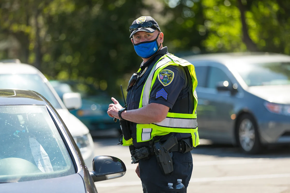 A police officer in a mask and safety vest stands near parked cars in a parking lot on a sunny day, prepared for Critical Threat Response.