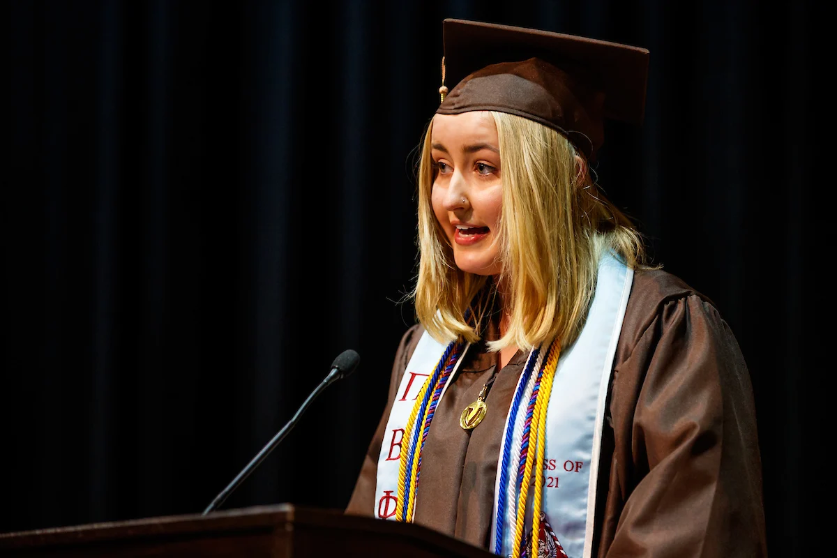 A student in a graduation cap and gown speaks at a podium during a ceremony where transcript services are available for graduates.