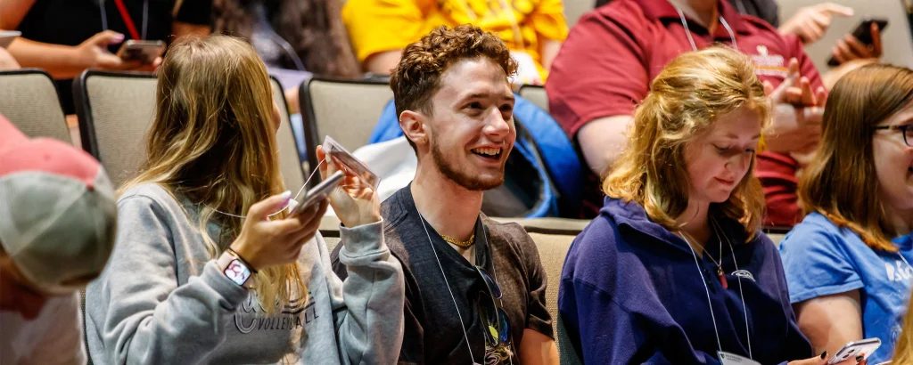 Three students sit in an auditorium. Two are smiling and talking, while one sits quietly with her FOCUS on her phone.