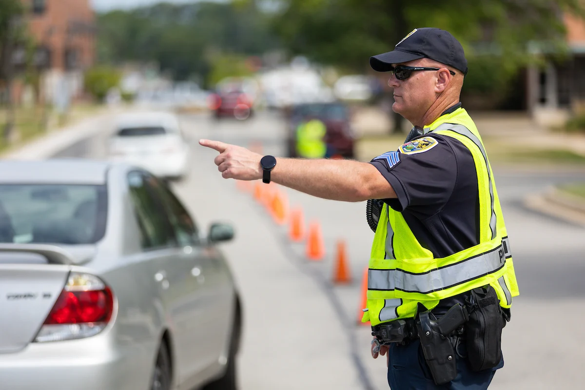 A Valparaiso University Police Department officer in a yellow vest directs traffic near orange cones on a city street.