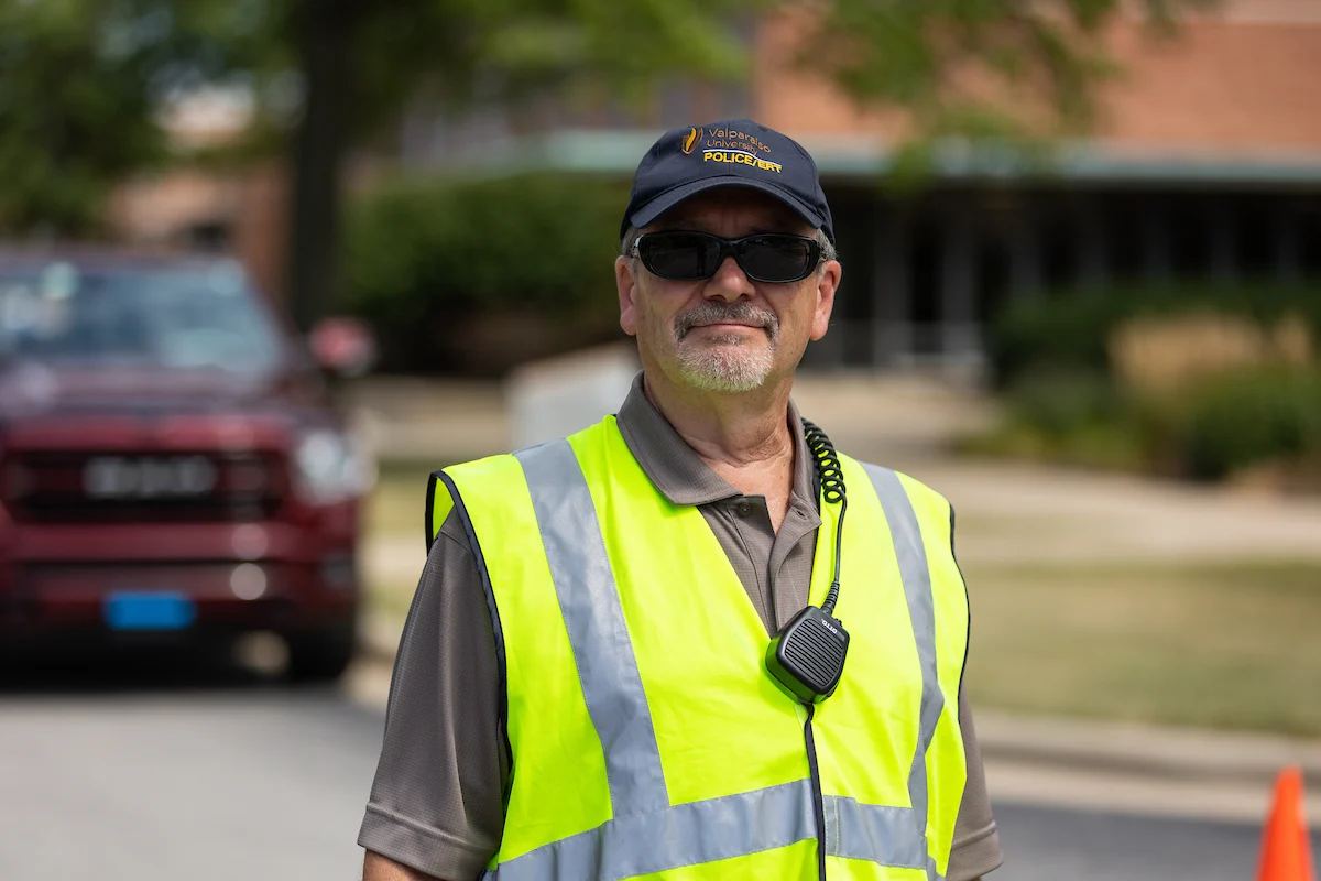 Man in a yellow safety vest and cap, wearing sunglasses and a radio, stands outdoors near a red vehicle, appearing ready for Critical Threat Response situations.
