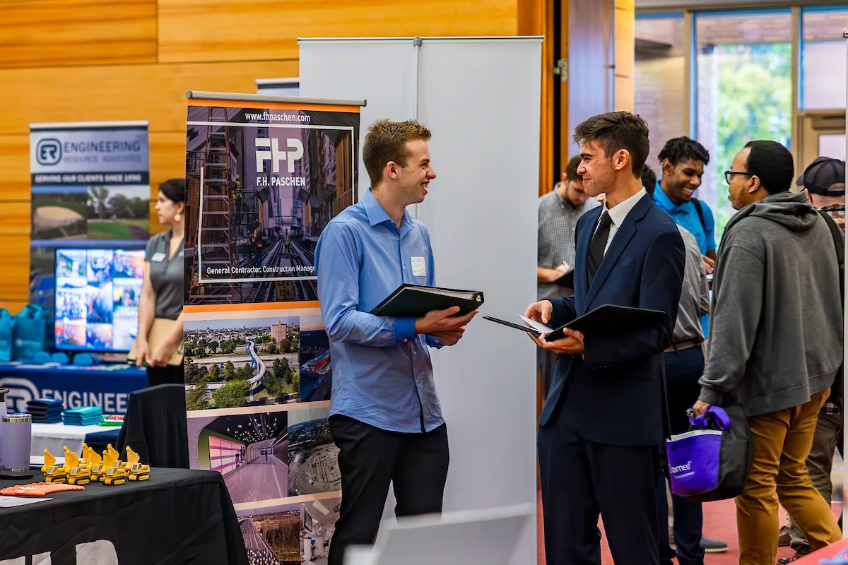 Two men talk at a Vale career fair booth with engineering posters and other attendees in the background.