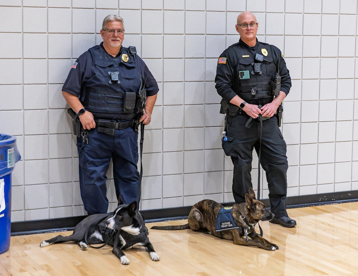Two Valparaiso University Police Department officers stand against a wall with two police dogs lying on the gym floor in front of them.