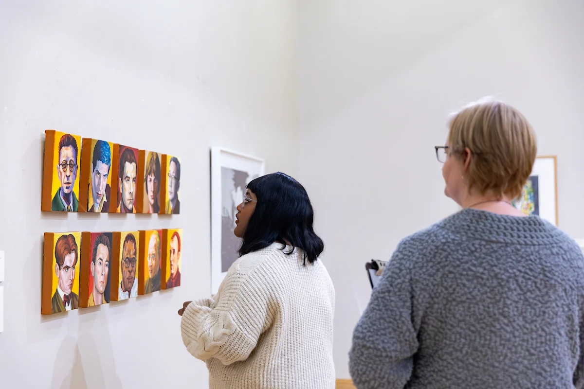 Two people admire colorful portrait paintings displayed on a white wall in an art gallery, immersed in the vibrant world of arts & culture.
