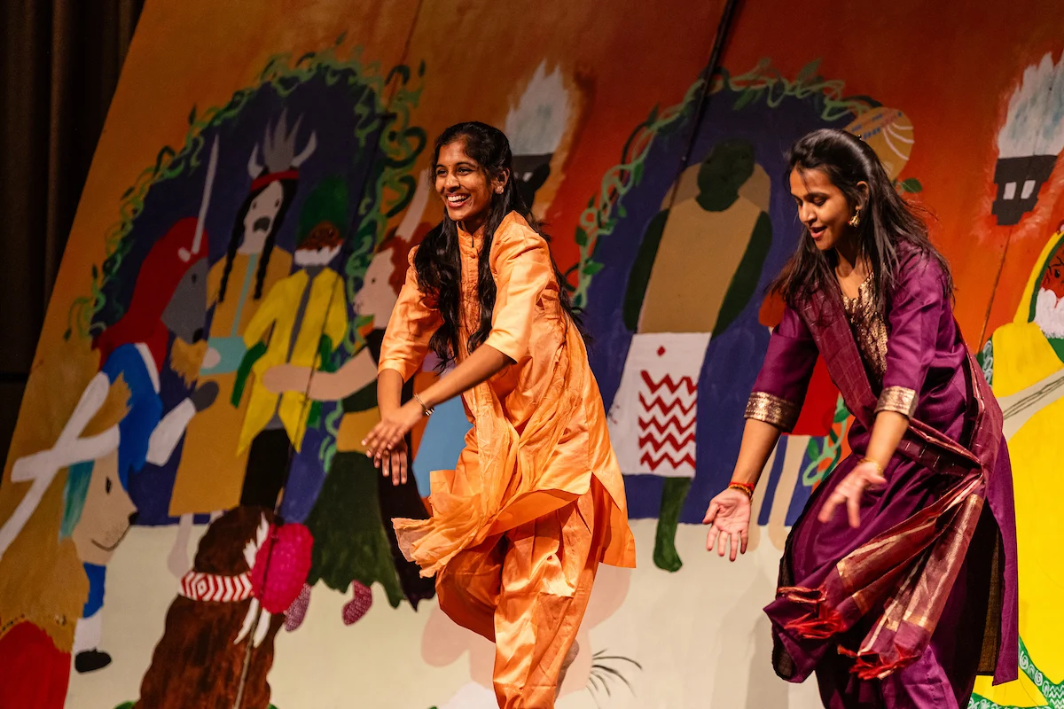Two girls in traditional Vale outfits dance joyfully on stage, with a colorful mural backdrop behind them.