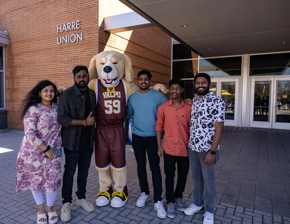 Five people stand smiling with a dog mascot in a sports jersey outside Harre Union, capturing the lively spirit of campus.