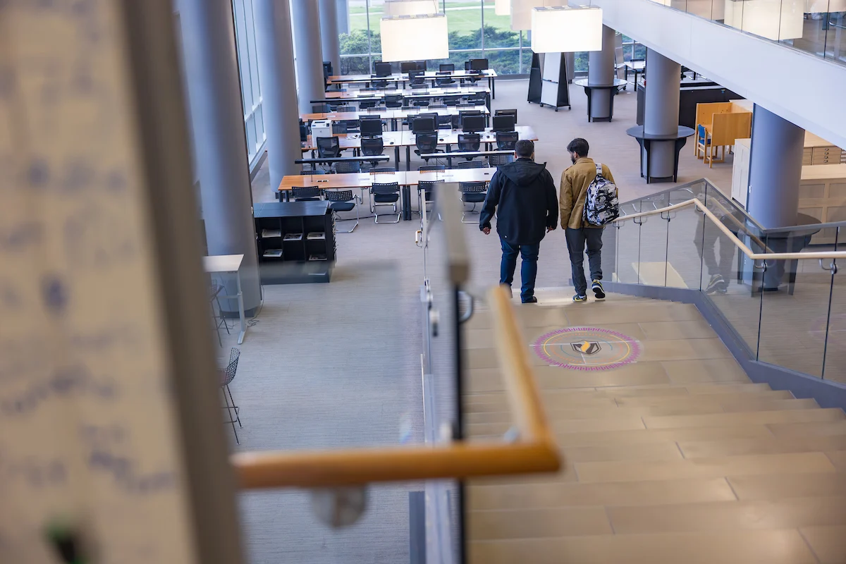 Two people walk down stairs into a modern, open campus study area with desks and large windows.