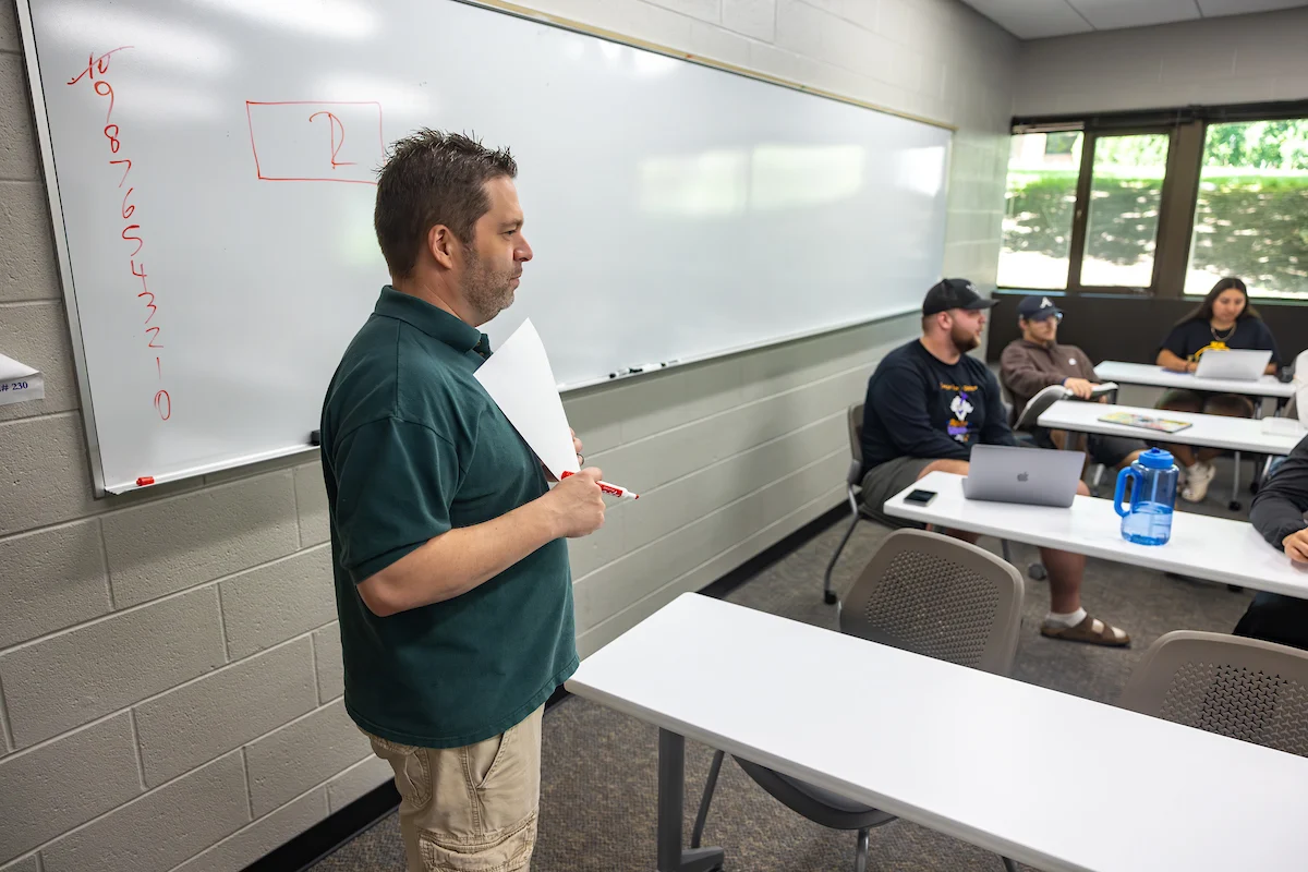 A teacher stands by a whiteboard while students sit at desks with laptops in a classroom, as the registrar organizes important academic records nearby.