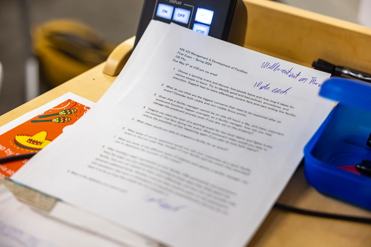 Close-up of a printed document with handwritten notes, possibly for the registrar, on a cluttered desk near a phone and a blue container.