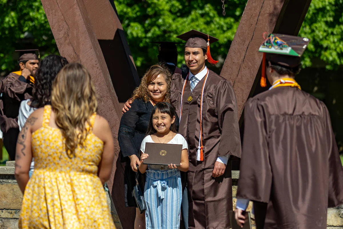 A graduate poses with family for a photo at an outdoor graduation ceremony on a sunny day, celebrating this milestone and sharing the moment with loved ones after receiving important Information for Graduates.